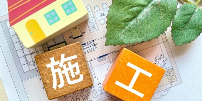 家の模型と「施工」の文字ブロック、間取り図と葉の装飾