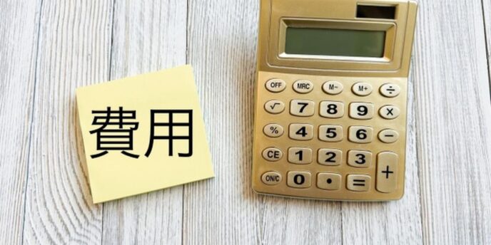 木目調の背景に置かれた電卓と「費用」と書かれたメモ用紙。費用計算のイメージ。