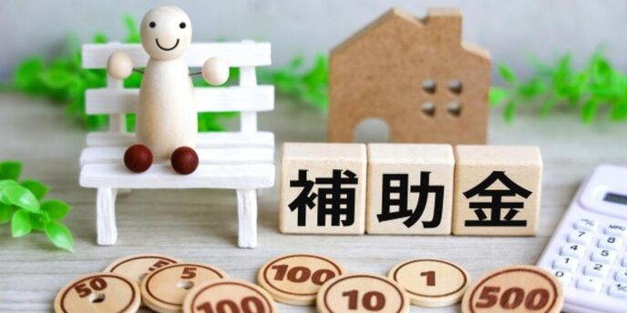 木製の家模型やコイン、ベンチに座る人形と「補助金」の文字が並ぶ温かみのある構成。
