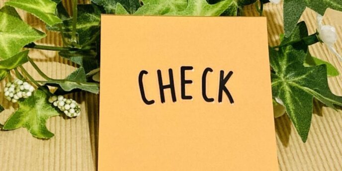 オレンジ色のメモに「CHECK」と書かれ、背景には観葉植物と白い花が添えられている構成。