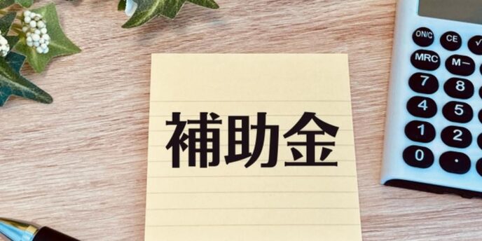 付箋に書かれた「補助金」の文字と電卓、ペン、観葉植物が並ぶデスクの上のイメージ。