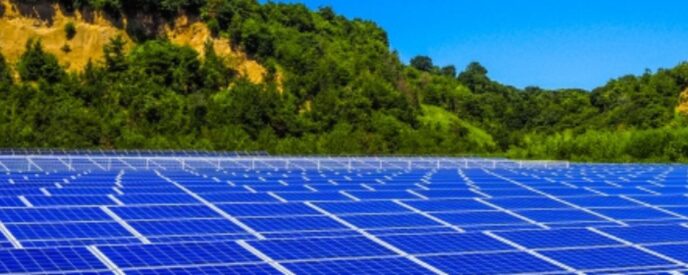 青空と緑の山を背景に、前景には多数の太陽光パネルが整然と並ぶ再生可能エネルギーの風景。