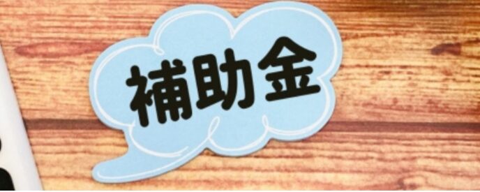 木目の机の上に「補助金」と書かれた吹き出し、電卓、ペン、観葉植物